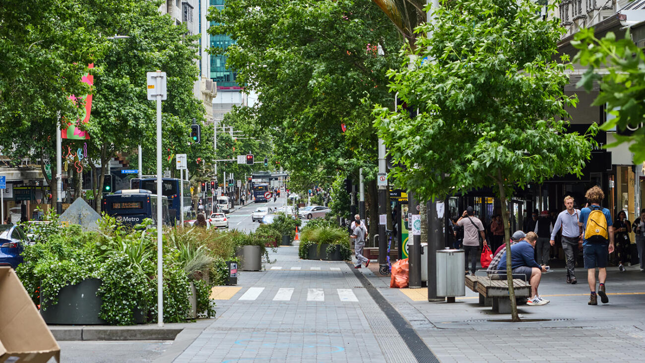 Tree-lined city street with wide footpaths, planting beds, public seating, and people walking by. 